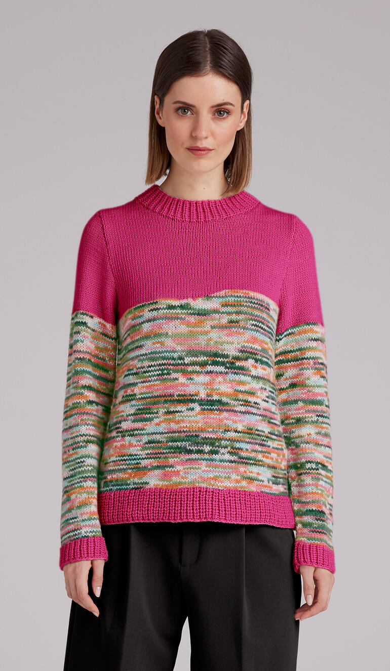 Frau trägt einen bunten Pullover mit geometrischem Muster, steht vor einem neutralen Hintergrund.