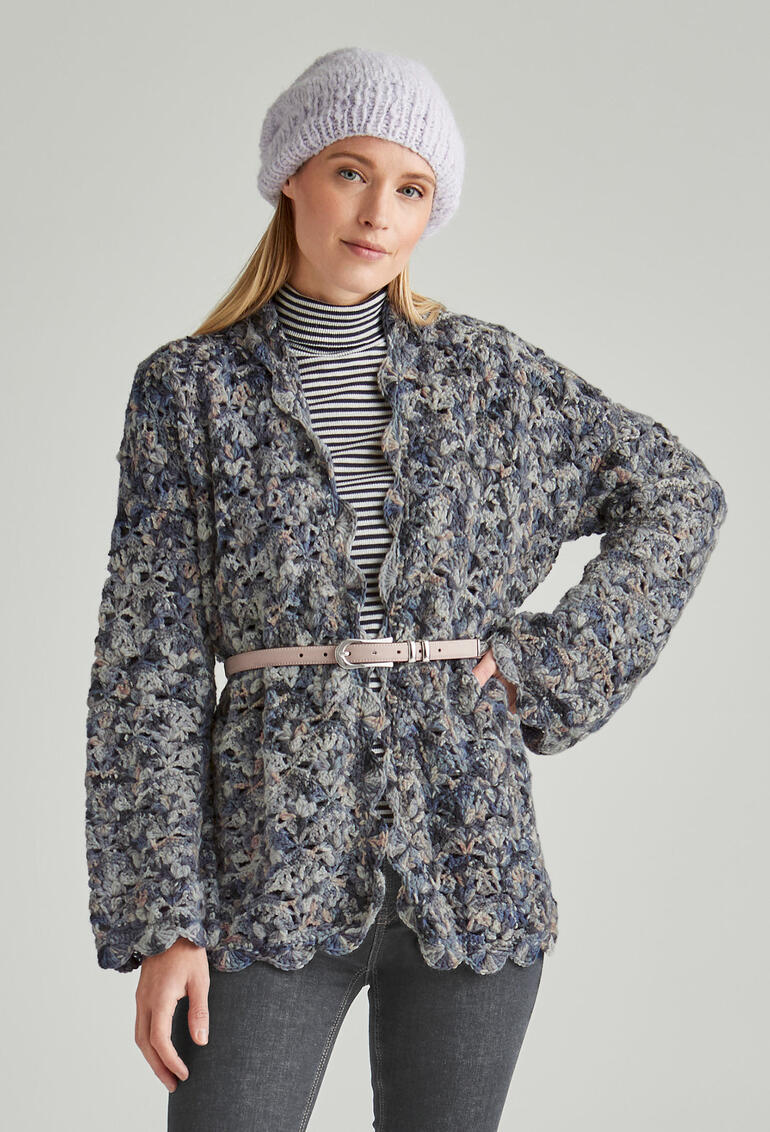 Frau in einer gehäkelten Jacke mit einem Gürtel, steht vor einem hellen Hintergrund.