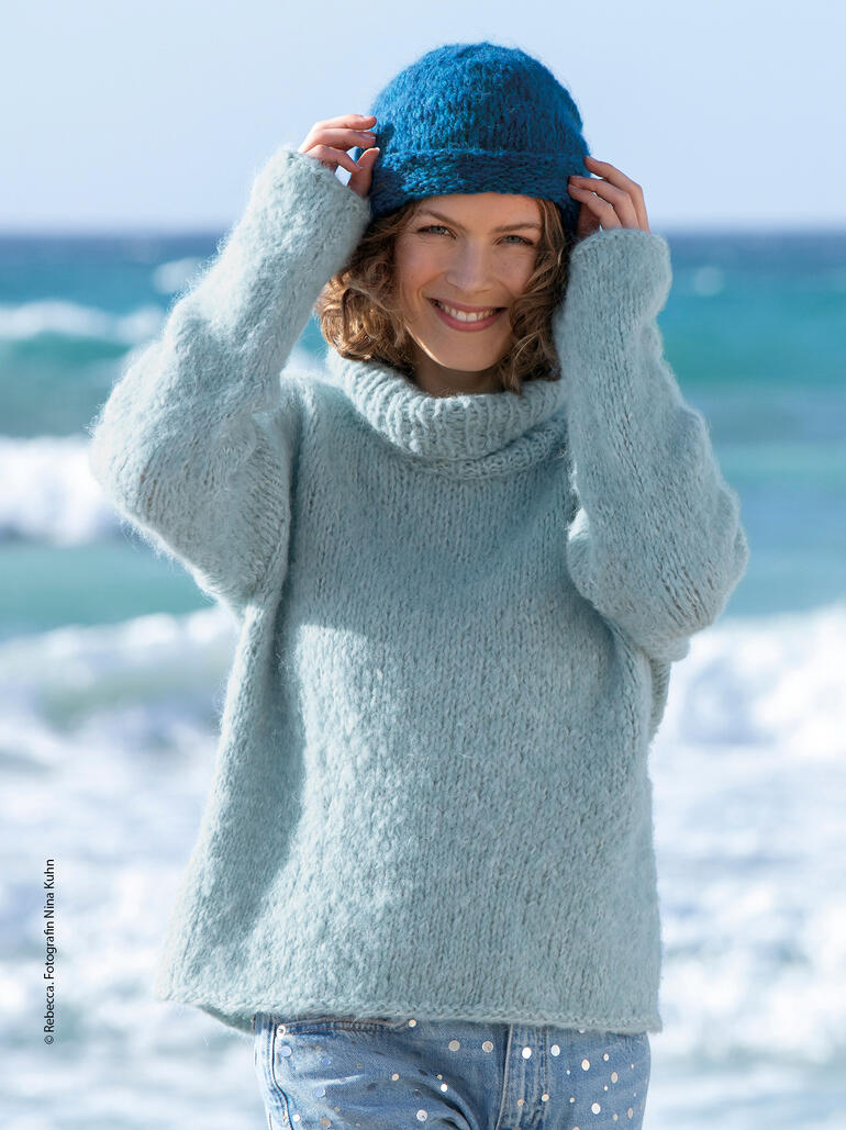 Frau in einem hellblauen Pullover mit hohem Kragen, trägt eine Mütze am Strand und lächelt.