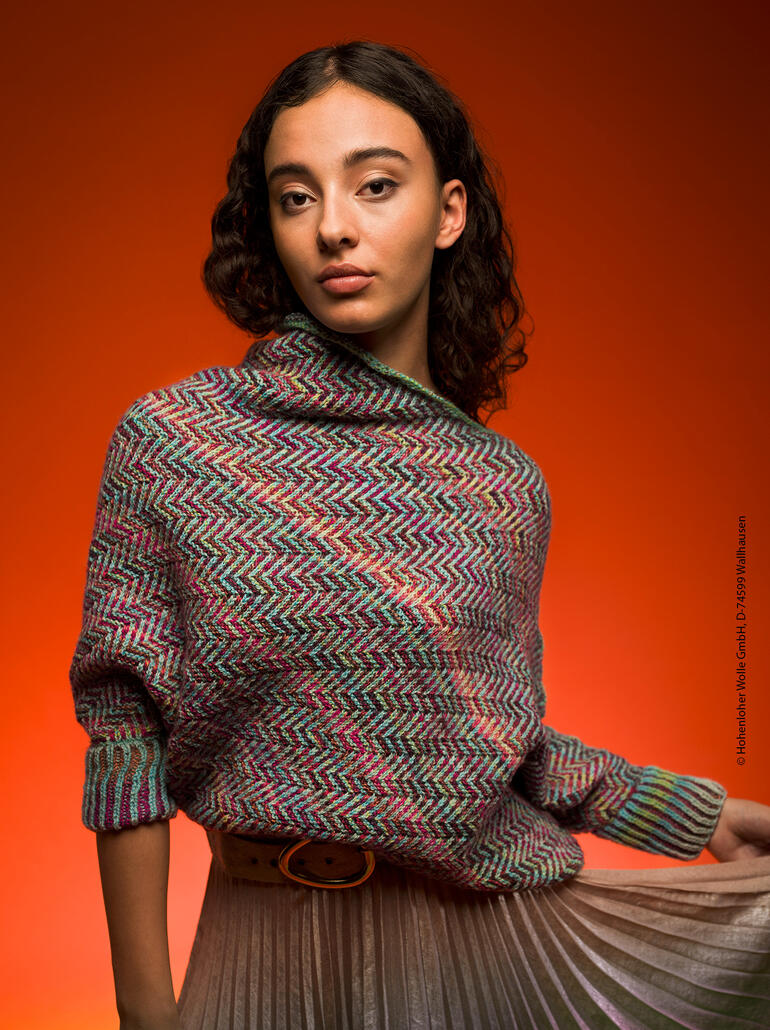 Frau trägt einen bunten Pullover mit Wellenmuster, steht vor einem orangefarbenen Hintergrund und schaut in die Kamera.