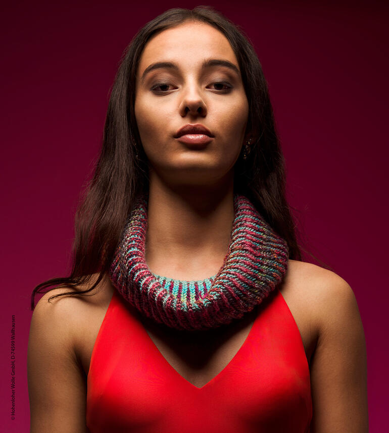 Frau trägt einen bunten Loop-Schal aus Strick, steht vor einem lila Hintergrund und sieht ernst aus.