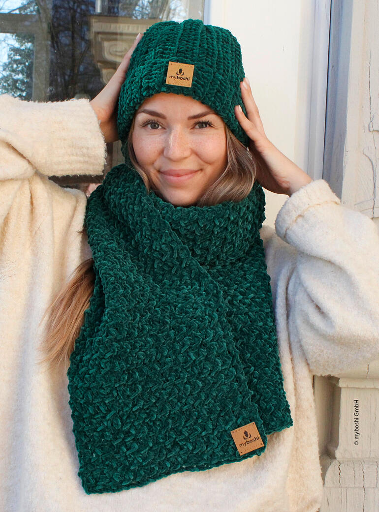 Frau mit Schal und Mütze in Dunkelgrün, aus Samt gehäkelt, lächelt in die Kamera.