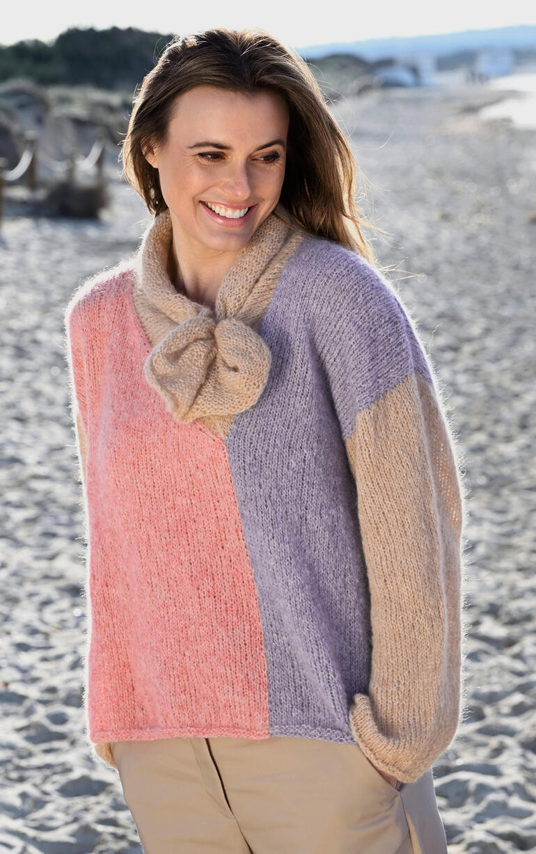 Frau in buntem Pullover aus Linie 550 Prato, mit einer Schleife, sitzt am Strand.