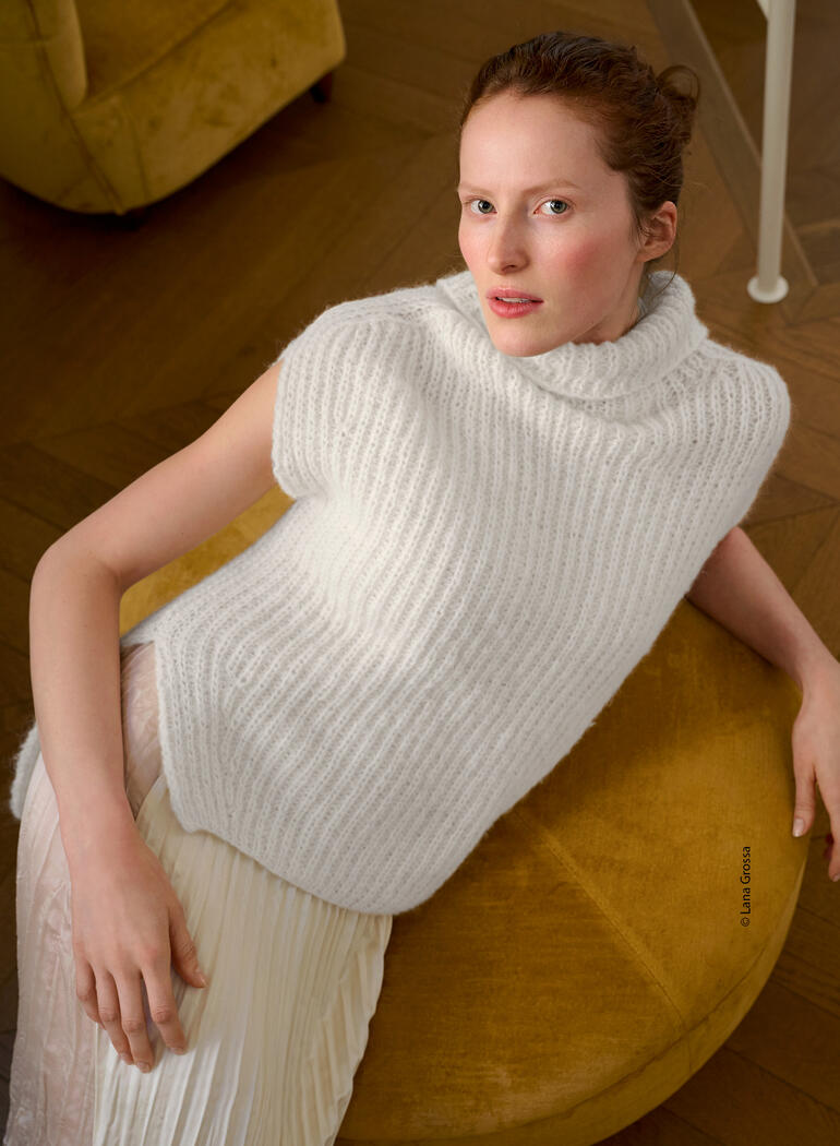 Frau trägt weißen Pullunder aus Nebbia, lächelt auf einem gelben Sessel.