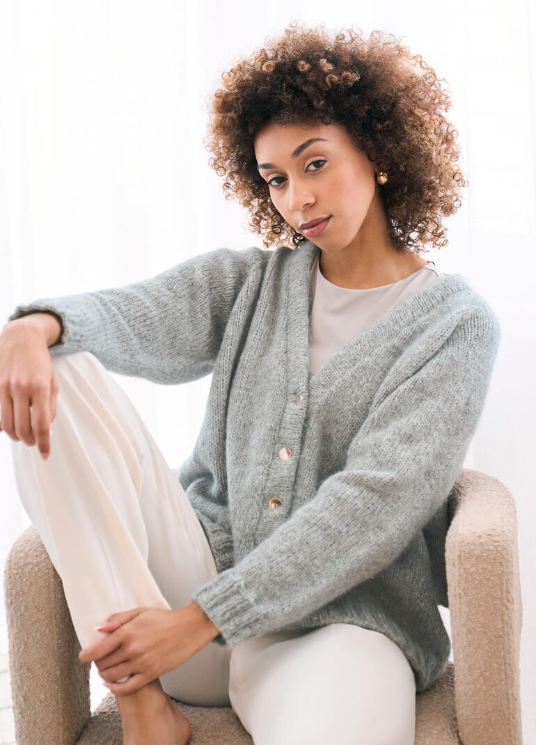 Frau in grauer Jacke aus Softest Alpaca, sitzt auf einem Sessel.