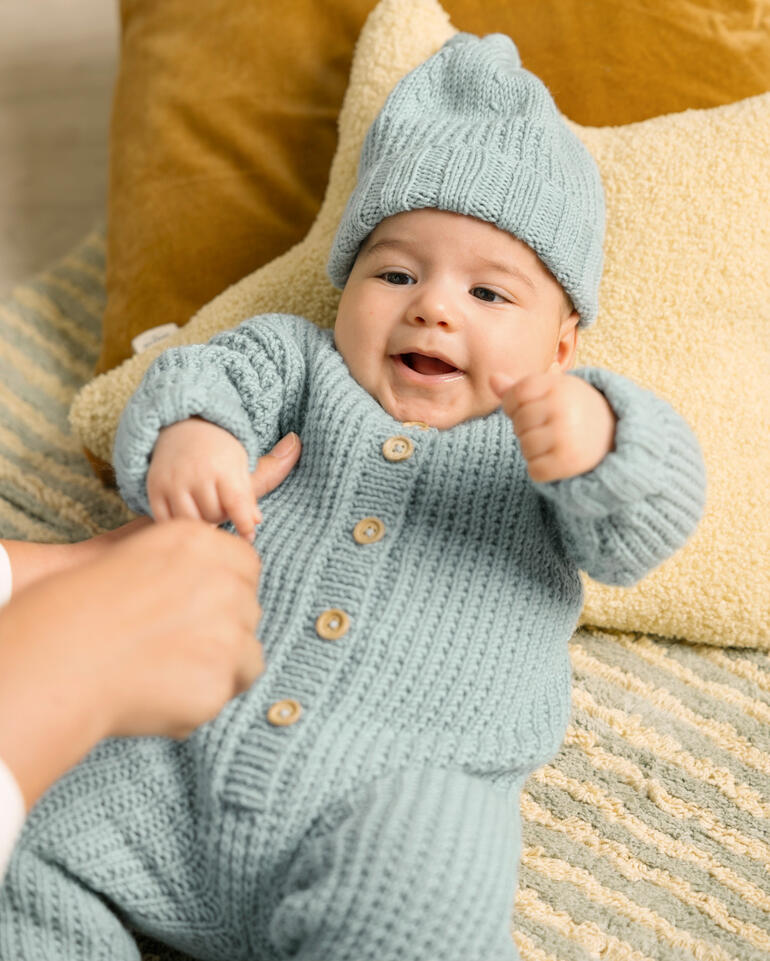Baby in hellblauem Strampler und Mütze, spielt auf einem Teppich.