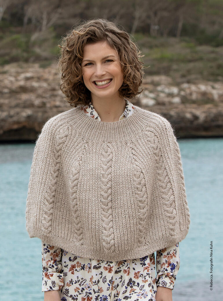 Lächelnde Frau in einem beigen Poncho aus dickem Garn vor einer Wasserfläche.
