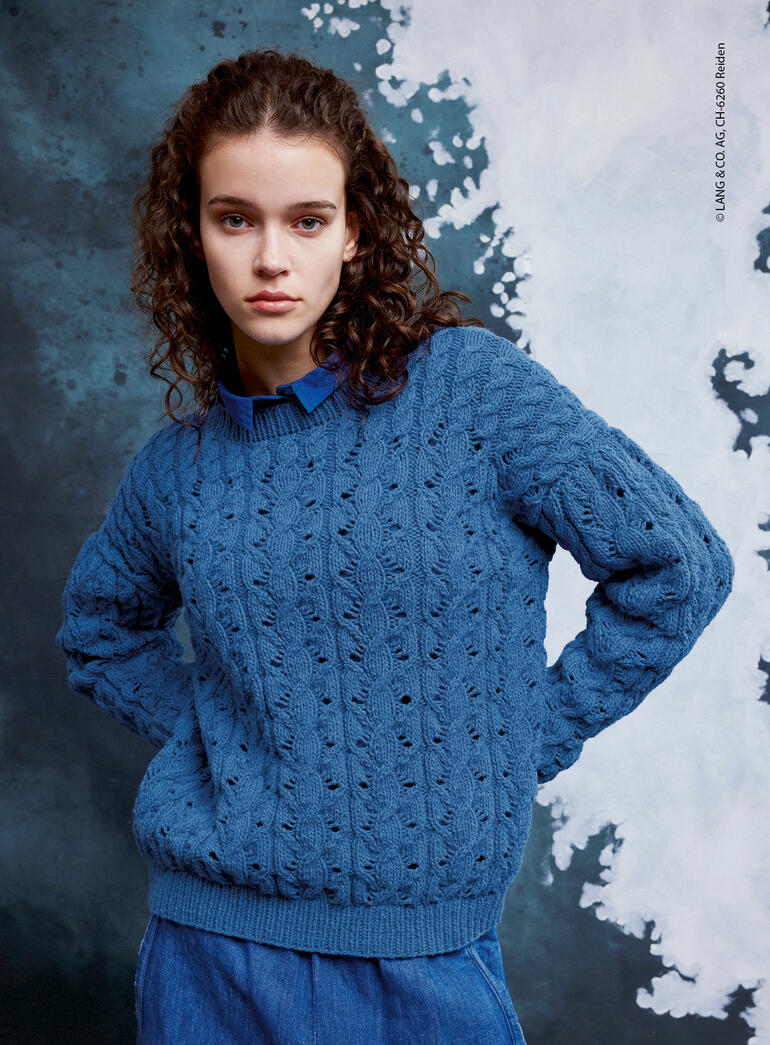 Frau trägt einen blauen Pullover mit Lochmuster, schaut direkt in die Kamera.