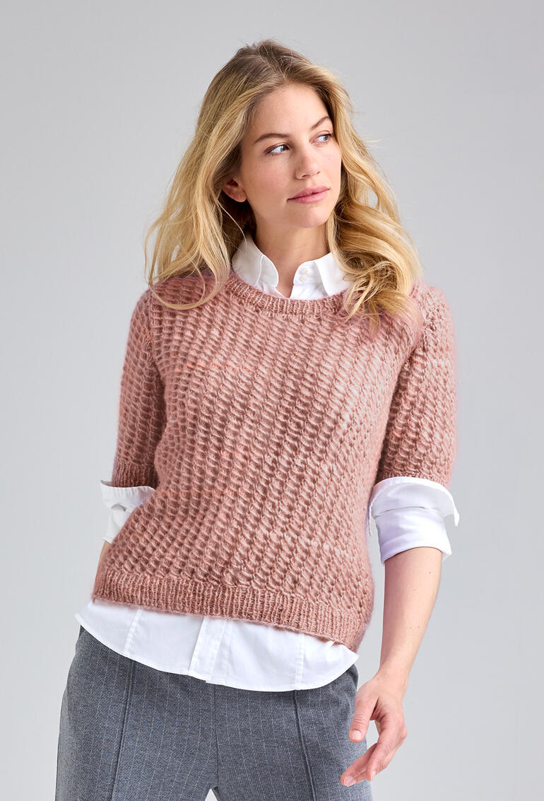 Frau trägt einen rosafarbenen Pullover mit Reliefmuster, schaut nachdenklich.