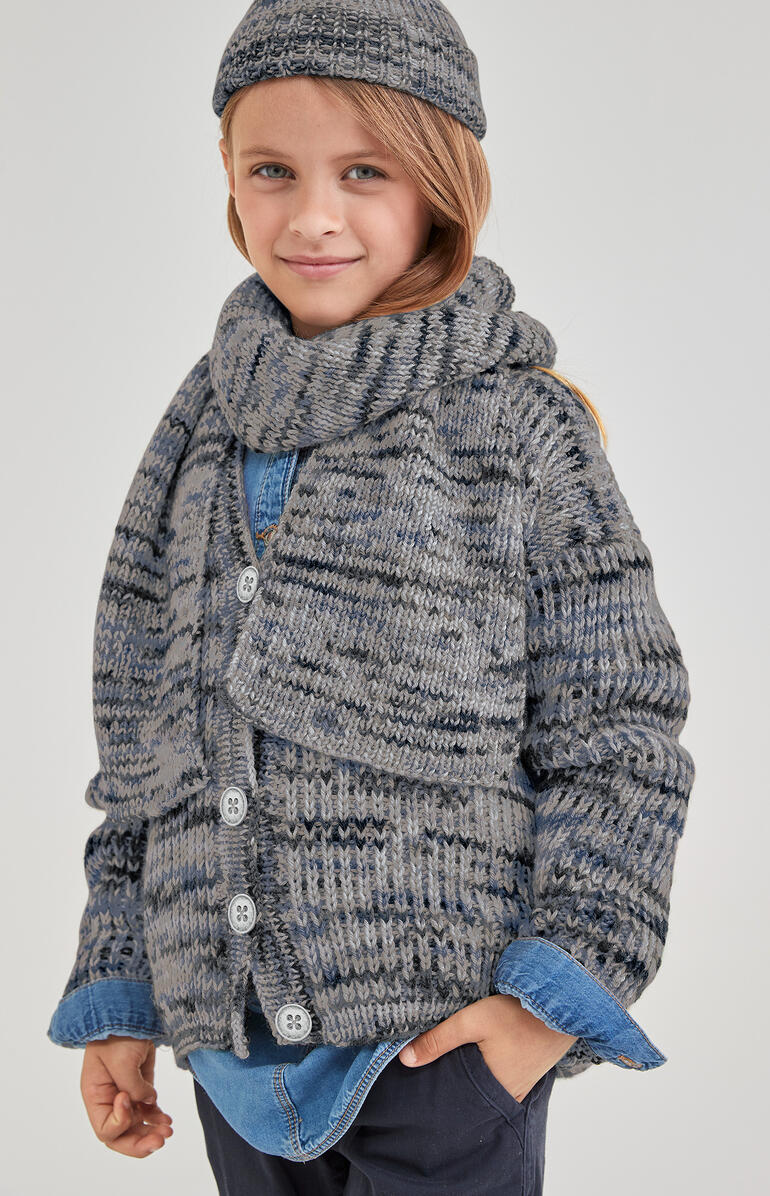 Junge in Cardigan aus Merino-Print mit Schal und Mütze, freundlich lächelnd.