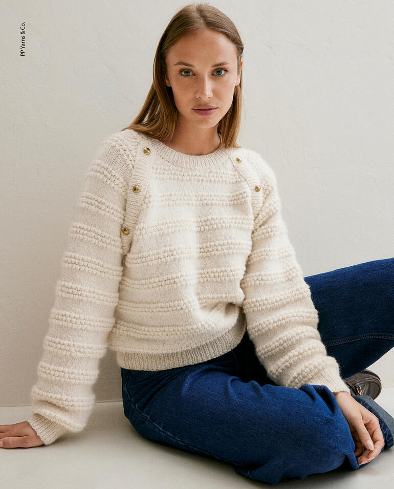 Frau sitzt in einem weißen Pullover aus Phil Baby Alpaga von Phildar.