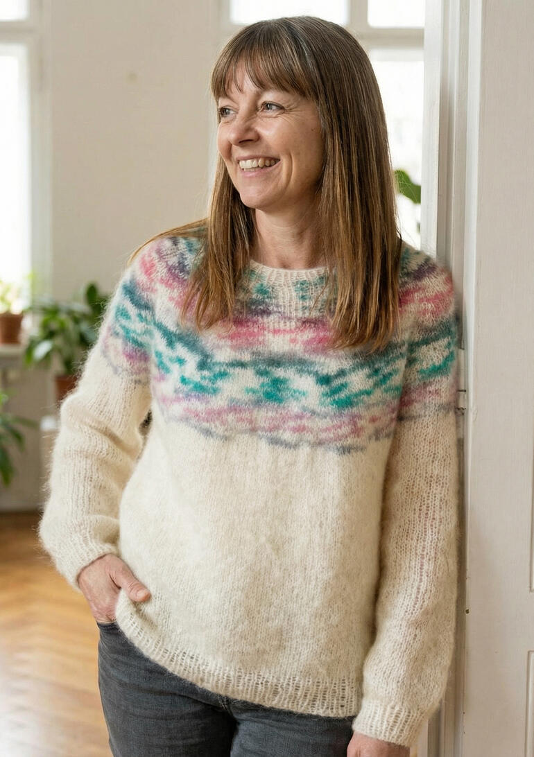 Frau in einem hellen Pullover mit buntem Muster, lächelt vor einem Fenster.
