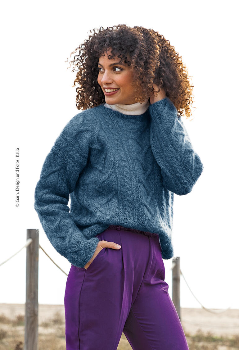 Blauer Pullover aus Ingenua, Modell mit Locken und lächelnd in der Natur.
