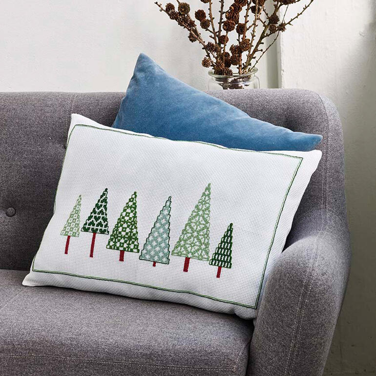 Kissenhülle mit verschiedenen Weihnachtsbaum-Motiven auf einer grauen Couch.
