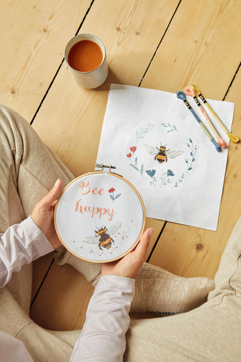 Person hält einen Stickrahmen mit dem Schriftzug 'Bee happy' und einem Blattdesign.