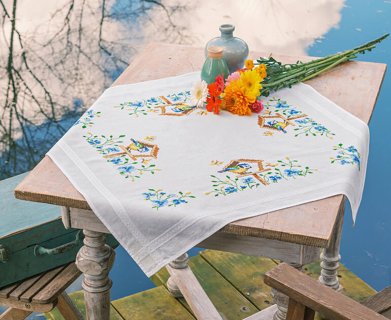 Tischdecke mit Vogelhäuschen-Motiven auf einem Tisch am Wasser, dekoriert mit Blumen