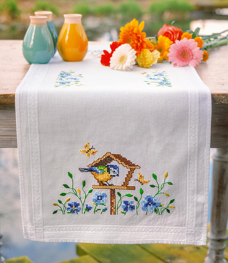 Tischläufer mit buntem Vogelhäuschen und Blumenarrangement auf einem Tisch.